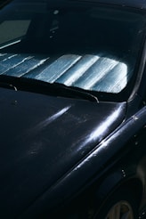 A close-up view of a car windshield with a reflective sunshade in place to block sunlight. The surface of the car is visible with reflections and subtle scratches on a dark paint.