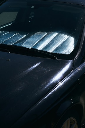 A close-up view of a car windshield with a reflective sunshade in place to block sunlight. The surface of the car is visible with reflections and subtle scratches on a dark paint.