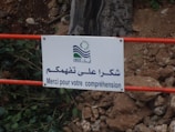 A white sign with a logo and multilingual text is attached to a red metal railing. The text reads in Arabic and French, expressing thanks for understanding. The background includes scattered rocks and vegetation.