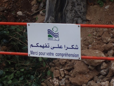 A white sign with a logo and multilingual text is attached to a red metal railing. The text reads in Arabic and French, expressing thanks for understanding. The background includes scattered rocks and vegetation.