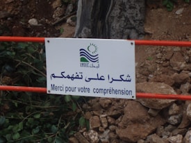 A white sign with a logo and multilingual text is attached to a red metal railing. The text reads in Arabic and French, expressing thanks for understanding. The background includes scattered rocks and vegetation.