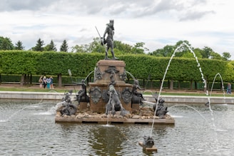A grand fountain featuring a large statue of a figure holding a staff, surrounded by smaller sculptures depicting mythical creatures and humans. Water jets elegantly arc into the air from the base of the fountain. Lush, green hedges and trees form the background, offering a serene and natural setting with a few people strolling nearby.