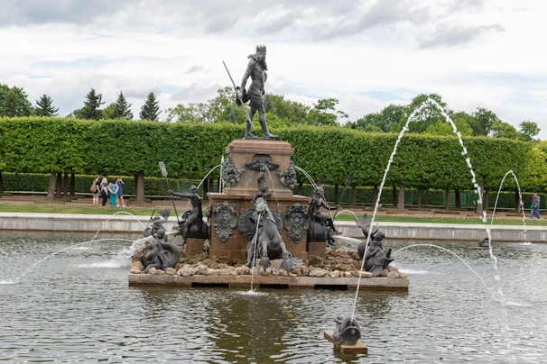 A grand fountain featuring a large statue of a figure holding a staff, surrounded by smaller sculptures depicting mythical creatures and humans. Water jets elegantly arc into the air from the base of the fountain. Lush, green hedges and trees form the background, offering a serene and natural setting with a few people strolling nearby.