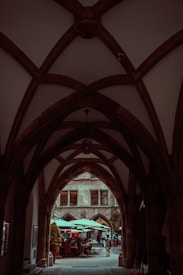 A gothic-style archway frames the view of an outdoor cafe area. People casually walk and sit under large umbrellas, creating a lively atmosphere. The stone architecture of the surrounding buildings adds a historical charm.