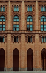 The facade of a historic building features ornate architectural designs with arched windows and intricate carvings. The structure is made of light brown brick, and the windows are tall with a distinctive arch at the top, incorporating decorative elements around the frames.