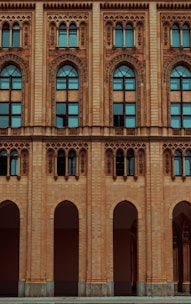 The facade of a historic building features ornate architectural designs with arched windows and intricate carvings. The structure is made of light brown brick, and the windows are tall with a distinctive arch at the top, incorporating decorative elements around the frames.