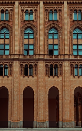 The facade of a historic building features ornate architectural designs with arched windows and intricate carvings. The structure is made of light brown brick, and the windows are tall with a distinctive arch at the top, incorporating decorative elements around the frames.