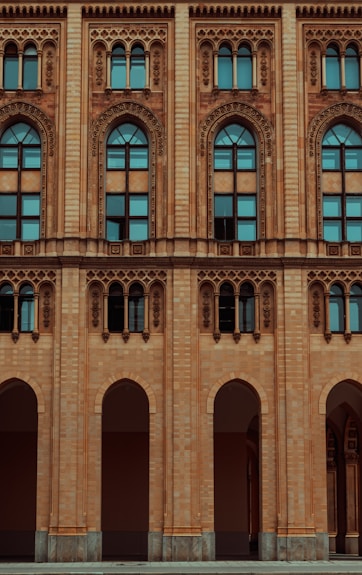 The facade of a historic building features ornate architectural designs with arched windows and intricate carvings. The structure is made of light brown brick, and the windows are tall with a distinctive arch at the top, incorporating decorative elements around the frames.