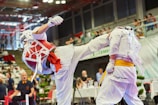 Two individuals are engaged in a martial arts competition. One person is executing a high kick while the other is defending. Both are wearing protective gear, including helmets, gloves, and body padding. The competition takes place in an indoor arena with an audience in the background.