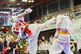 Two individuals are engaged in a martial arts competition. One person is executing a high kick while the other is defending. Both are wearing protective gear, including helmets, gloves, and body padding. The competition takes place in an indoor arena with an audience in the background.