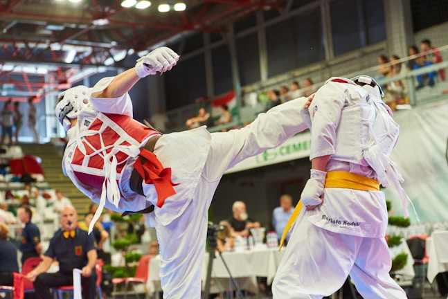 Two individuals are engaged in a martial arts competition. One person is executing a high kick while the other is defending. Both are wearing protective gear, including helmets, gloves, and body padding. The competition takes place in an indoor arena with an audience in the background.