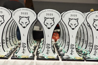 A collection of trophies with a stylized wolf head logo and the text 'European Championships 2023' displayed prominently. The trophies are arranged in neat rows, with a focus on the unique design and inscriptions in a foreign script. Each trophy is placed on a black base, and there are ribbons and medals attached at the bottom.