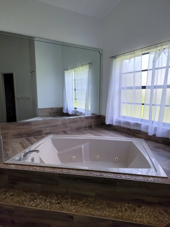 A corner bathtub is set into a raised wooden platform with a mosaic tile design lining the base. Large windows with sheer white curtains allow natural light to fill the room, reflecting softly on the tiled and wood surfaces. The atmosphere suggests a sense of tranquility and calm.