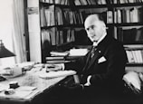 A man in formal attire is seated at a desk cluttered with papers and books, surrounded by bookshelves filled with more books. The setting appears to be an office or study, with ample natural light coming through a window. He has a serious, contemplative expression, with a hand poised over a stack of documents.