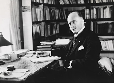 A man in formal attire is seated at a desk cluttered with papers and books, surrounded by bookshelves filled with more books. The setting appears to be an office or study, with ample natural light coming through a window. He has a serious, contemplative expression, with a hand poised over a stack of documents.