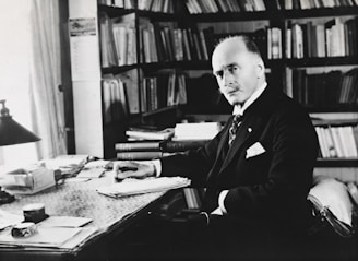 A man in formal attire is seated at a desk cluttered with papers and books, surrounded by bookshelves filled with more books. The setting appears to be an office or study, with ample natural light coming through a window. He has a serious, contemplative expression, with a hand poised over a stack of documents.