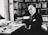 A man in formal attire is seated at a desk cluttered with papers and books, surrounded by bookshelves filled with more books. The setting appears to be an office or study, with ample natural light coming through a window. He has a serious, contemplative expression, with a hand poised over a stack of documents.