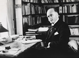 A man in formal attire is seated at a desk cluttered with papers and books, surrounded by bookshelves filled with more books. The setting appears to be an office or study, with ample natural light coming through a window. He has a serious, contemplative expression, with a hand poised over a stack of documents.