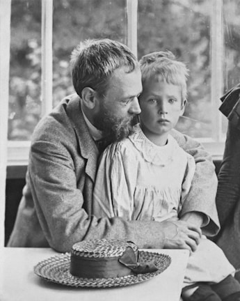 A black and white photograph depicts an adult male with a beard holding a young child on his lap. The man is wearing a suit jacket and appears to be comforting or protecting the child, who is dressed in a light-colored, old-fashioned clothing. A straw hat with a ribbon rests on a table in the foreground. The background shows a window with a blurred view of foliage outside.