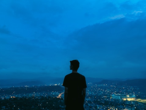A cinematic still showing a silhouetted hero standing atop a city skyline under a stormy sky.