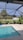 An outdoor pool with clear blue water is surrounded by a screened enclosure. The area is landscaped with potted plants and there is a lush green lawn with several palm trees visible in the background. A white building is situated beyond the lawn, and the sky above is clear and sunny.