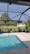 An outdoor pool with clear blue water is surrounded by a screened enclosure. The area is landscaped with potted plants and there is a lush green lawn with several palm trees visible in the background. A white building is situated beyond the lawn, and the sky above is clear and sunny.