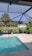 An outdoor pool with clear blue water is surrounded by a screened enclosure. The area is landscaped with potted plants and there is a lush green lawn with several palm trees visible in the background. A white building is situated beyond the lawn, and the sky above is clear and sunny.