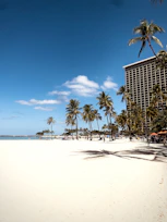 A sandy beach stretches along the coastline with tall palm trees lining the area. A large hotel building is seen to the right, and there are people walking and relaxing near the water. The sky is clear with a few clouds, and the sun casts long shadows on the sand.