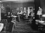 Several people are seated in a cozy, vintage-style reading room, engaged in reading books and magazines. The room is decorated with framed portraits on the walls, potted plants, and wicker furniture. A white statue is displayed prominently on the right side of the room, and a bookshelf filled with books is visible in the background.