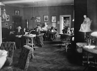 Several people are seated in a cozy, vintage-style reading room, engaged in reading books and magazines. The room is decorated with framed portraits on the walls, potted plants, and wicker furniture. A white statue is displayed prominently on the right side of the room, and a bookshelf filled with books is visible in the background.