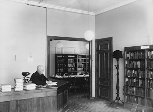 A man is sitting at a wooden desk surrounded by stacks of books in a library setting. The room features wooden bookshelves filled with books, and a sign labeled 'Psykoteknikk'. Decorative ceiling lights hang from above, and there are wooden doors and a coat stand nearby.