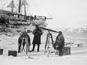 Three people dressed in heavy fur coats are engaged in an activity on a snowy landscape. They appear to be conducting an observation with a telescope or similar instrument mounted on tripods. A large ship, partially covered in snow, is visible in the background, with ice and mountains in the distance.