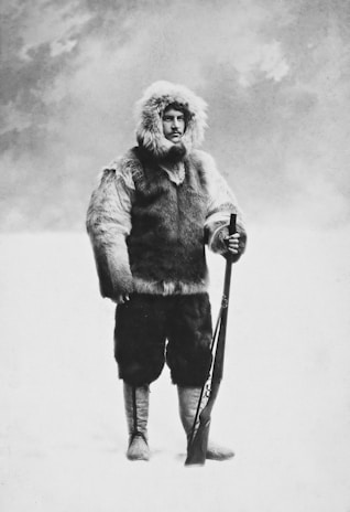 A person is dressed in a fur coat, hood, and boots, holding a rifle. The clothing appears thick and suited for cold environments. The background is a snowy or misty landscape, creating a stark contrast with the figure.