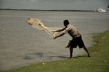A person is casting a fishing net into a large body of water from a grassy shoreline. In the background, calm waters extend towards a distant horizon with a few small boats and a glimpse of shoreline. The sky above is overcast, adding a muted tone to the scene.