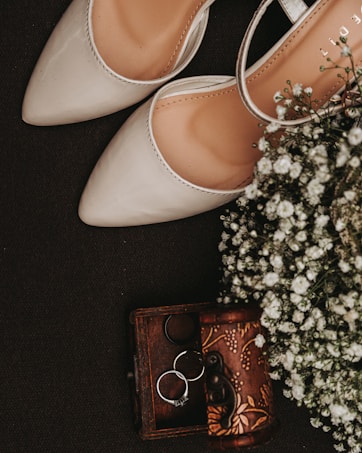 A pair of elegant beige high-heeled shoes placed next to a small vintage wooden box containing a few metal rings. Delicate white flowers complement the arrangement, adding a touch of natural elegance.