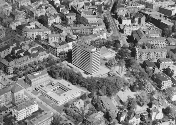 An aerial view of a cityscape featuring a large rectangular office building at the center. The surrounding area includes a mix of residential and commercial buildings, tree-lined streets, and open green spaces. The architecture is varied, with both older brick structures and more modern constructions. Roads intersect at various points, and there is visible urban planning with circular intersections and organized blocks.