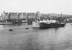 A historical black and white photograph of a harbor scene with ships anchored and a large building in the background. The water stretches across the foreground with several boats visible. The architecture of the building is distinctive with pointed roof sections and multiple windows. City structures and hills can be seen in the distance.