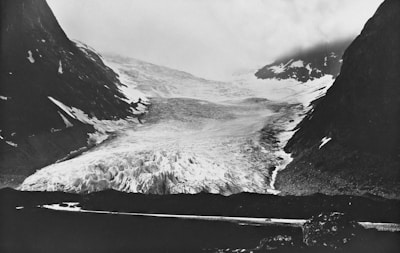 A vast glacier is nestled between two dark, rocky mountain slopes under an overcast sky. The surface of the glacier appears rugged and uneven, composed of ice and snow, with some visible crevasses. The surrounding mountains are steep, with traces of snow along their sides. The overall scene is misty, with low-hanging clouds obscuring the view further back.