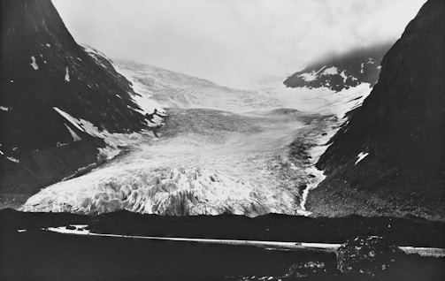 A vast glacier is nestled between two dark, rocky mountain slopes under an overcast sky. The surface of the glacier appears rugged and uneven, composed of ice and snow, with some visible crevasses. The surrounding mountains are steep, with traces of snow along their sides. The overall scene is misty, with low-hanging clouds obscuring the view further back.
