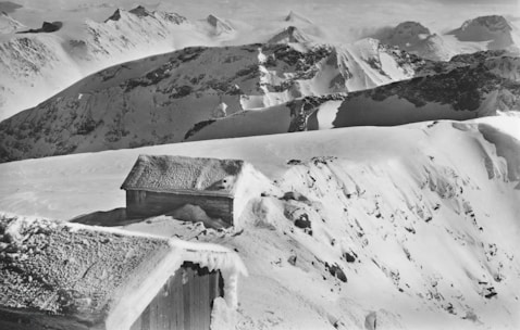 A prefabricated cottage nestled on a steep mountain slope with snow-capped peaks in the background.
