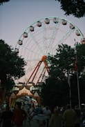 A large Ferris wheel towers over a park entrance with a sign that reads 'Altın Park.' The structure is illuminated with lights, creating a striking contrast against the twilight sky. People are walking and gathering at the entrance, adding a lively atmosphere. The scene is framed by trees, and various banners and flags can be seen.