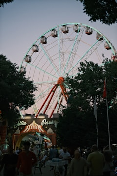A large Ferris wheel towers over a park entrance with a sign that reads 'Altın Park.' The structure is illuminated with lights, creating a striking contrast against the twilight sky. People are walking and gathering at the entrance, adding a lively atmosphere. The scene is framed by trees, and various banners and flags can be seen.