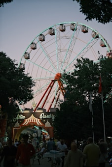 A large Ferris wheel towers over a park entrance with a sign that reads 'Altın Park.' The structure is illuminated with lights, creating a striking contrast against the twilight sky. People are walking and gathering at the entrance, adding a lively atmosphere. The scene is framed by trees, and various banners and flags can be seen.