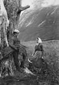 A black and white photograph depicts a rural scene with two people in traditional attire. A young man leans against a large tree, holding a bundle of grass, while a woman stands nearby with a scythe in her hand. The backdrop features a dramatic landscape of mountains and fields under a cloudy sky.