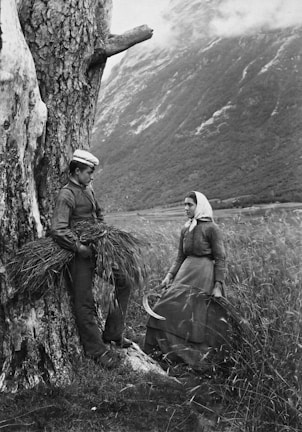 A black and white photograph depicts a rural scene with two people in traditional attire. A young man leans against a large tree, holding a bundle of grass, while a woman stands nearby with a scythe in her hand. The backdrop features a dramatic landscape of mountains and fields under a cloudy sky.