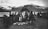 A group of people stand around a tent made of natural materials on grassy terrain near a body of water. Snow-capped mountains are visible in the background, and several individuals are dressed in traditional clothing. The setting appears to be a gathering or market as items are laid out on the ground.