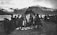 A group of people stand around a tent made of natural materials on grassy terrain near a body of water. Snow-capped mountains are visible in the background, and several individuals are dressed in traditional clothing. The setting appears to be a gathering or market as items are laid out on the ground.