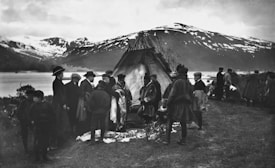 A group of people stand around a tent made of natural materials on grassy terrain near a body of water. Snow-capped mountains are visible in the background, and several individuals are dressed in traditional clothing. The setting appears to be a gathering or market as items are laid out on the ground.