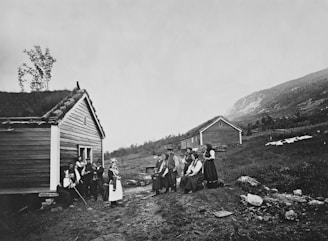 A group of people dressed in traditional attire are gathered outside two wooden houses in a rural setting. The environment features grassy hills and rocky terrain, with some trees and distant mountains. The houses have roofs covered with grass.