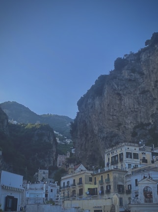A picturesque coastal village is nestled against a backdrop of steep, rocky cliffs. The sunlight casts a warm glow on the cluster of charming buildings, which are characterized by their light-colored facades and distinctive Mediterranean architecture. The sky above is clear and blue, enhancing the scenic beauty of the setting.